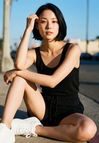 Noor naine mustas topis ja lühikestes pükstes, istub jalad risti tänavaserva peal päikese käes, vaatab otse kaamerasse.