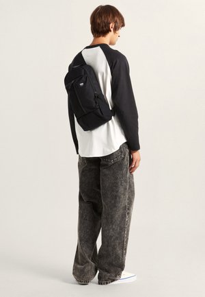 Black backpack with a front zipper pocket, attached to a person wearing a black and white shirt and loose gray jeans, standing from the back.