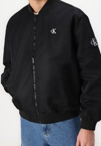 Bomber jacket nera con zip frontale, polsini e colletto a maglia. Presenta logo sul petto e tasca con chiusura a scatto. Tessuto liscio e lucido.