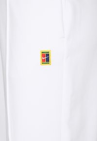 Witte broek met een kleurrijk geborduurd logo met een rechthoekige omranding en vierkante kleurvlakken, die de levendige ontwerpelementen benadrukt.