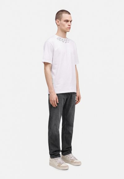 Jeune homme debout, portant un T-shirt blanc à manches courtes avec un lettrage près du col, un jean foncé, et des baskets beiges avec des lacets à motifs.
