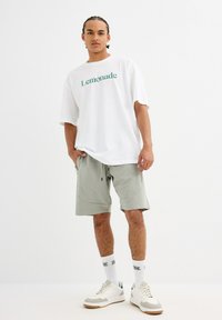 Una camiseta blanca con el texto "Limonada" en verde, pantalones cortos gris claro y zapatillas blancas con detalles en gris y marrón. Los calcetines presentan un diseño de logo.