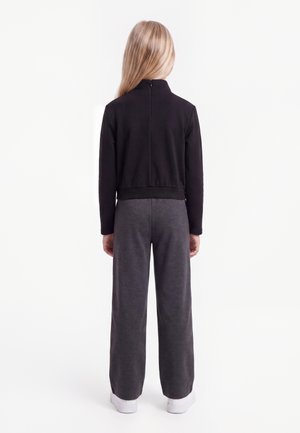 Kind mit langen blonden Haaren steht mit dem Rücken zur Kamera, trägt eine schwarze Jacke mit Reißverschluss, dunkelgraue Hose und weiße Turnschuhe auf weißem Hintergrund.