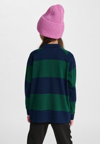 Stickad rosa mössa med ribbad textur, tillsammans med en långärmad, marinblå och grönrandig tröja. Svarta byxor syns nedanför.