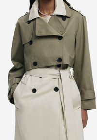 Gabardina corta verde oliva con botones negros, amplios solapas y un cinturón beige. Presenta dos bolsillos laterales y una textura suave.