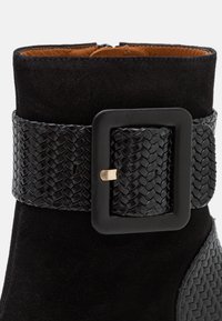 Bottine en daim noir avec bride à boucle en cuir tressé. Caractéristiques : texture lisse, boucle carrée et silhouette épurée.