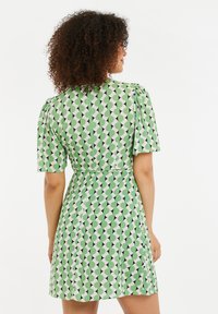 Vihreä lyhyt mekko, jossa on toistuva geometrinen kuvio, joka koostuu vihreästä, mustasta ja valkoisesta. Puffihihat ja istuva vyötärö korostavat muotoilua.