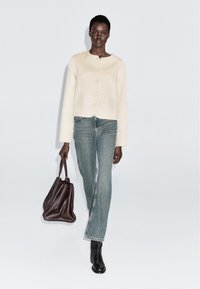 Vrouw die in een crèmekleurige knoopjas, blauwe jeans en zwarte laarzen loopt, en een grote, getextureerde donkerbruine handtas draagt tegen een effen achtergrond.