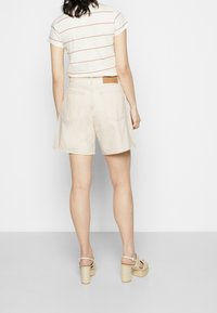 Pantalones cortos de algodón beige con una cintura alta, dos bolsillos traseros y dobladillos enrollados. Combinados con una blusa de manga corta a rayas y sandalias de tacón bloque en tono marrón.