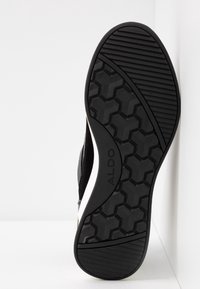 Semelle de chaussure noire avec motifs géométriques et rayés, marquée "ALDO" au centre, affichée verticalement sur fond blanc.