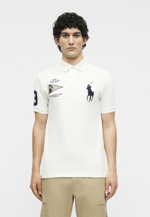 Άνδρας που φοράει λευκό πόλο μπλουζάκι με κεντημένο πατς ναυτικού ομίλου, λογότυπο polo σε θαλασσί χρώμα και μπεζ παντελόνι cargo, στέκεται μπροστά από απλό φόντο.