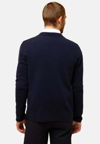 Marinblå tröja med strukturerad stickning, långa ärmar och V-ringning, sedd bakifrån. Rundad nederkant och minimal söm-detaljer.