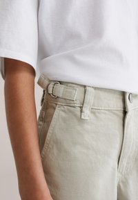 Nærbillede af maveparti iført lysebeige bukser med justerbar sidelukning og hvid kortærmet skjorte.