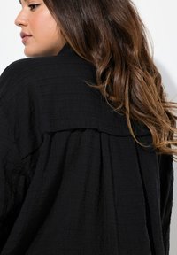 Black textured fabric shirt with a relaxed fit, featuring a back pleat and a structured collar. Soft, lightweight material.