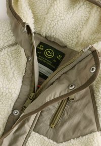 Fleece-gefütterte Jacke mit einem neutralen beigen Außenmaterial, mit Reißverschlusstaschen, Druckknopfverschluss und einem Pflegeetikett, das ein Smiley-Gesicht und Text zeigt.