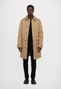 Beige padded coat with a collar, large buttons, side pockets, and a straight cut, worn over a black shirt and trousers. Black shoes.