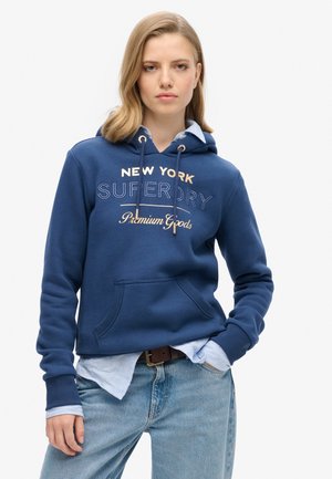Marineblauwe capuchontrui met geborduurde tekst, trekkoorden en een voorkeertas. Gelaagd over een lichtblauw overhemd met kraag.