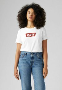 Camiseta de algodón blanca con un logo rojo de Levi's, combinada con unos jeans azules de talle alto. Escote sencillo y mangas cortas, con un corte casual.