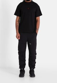Černé bavlněné tričko s malým červeným logem. V kombinaci s černými cargo kalhotami s několika kapsami a texturovanými detaily. Černé tenisky.