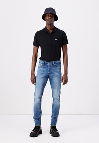 Cămașă polo neagră, blugi de culoare albastru deschis, îmbrăcați slim-fit, și pantofi chunky negri. Modelul poartă o pălărie gri tip bucket. Material din bumbac, design casual.