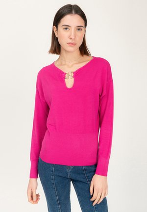 Maglione lavorato a maglia fucsia con scollatura a V e dettaglio dorato, polsini a costine e fondo aderente. Indossato con jeans blu.