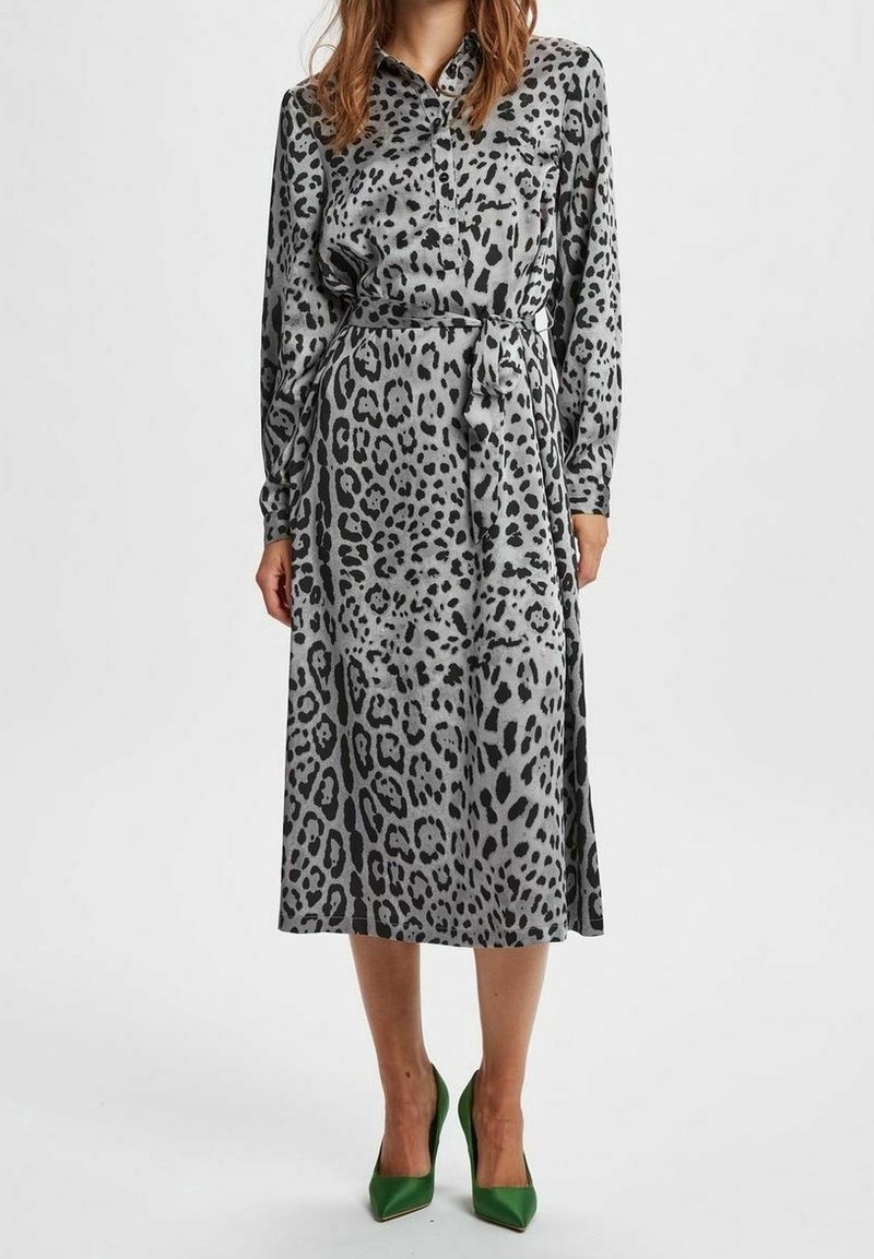 Femme portant une robe midi grise à manches longues ceinturée avec imprimé animalier noir, associée à des escarpins verts à bout pointu, debout sur fond blanc.