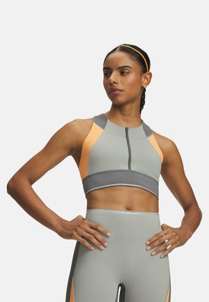 Athletische Frau mit geflochtenen Haaren in einem grauen und orangen Zip-Sport-BH und passenden hoch taillierten Leggings, Hände in die Hüften gestützt, schaut zur Seite.
