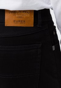 Primer plano del bolsillo trasero de unos jeans negros con una etiqueta de cuero marrón que dice "Eurex by Brax Established 1995" y parte de una camisa blanca metida.