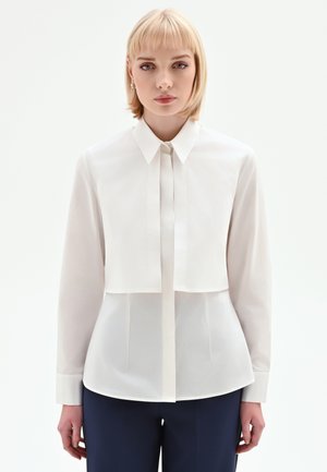 Camicia bianca con colletto, maniche lunghe e design a strati. Tessuto liscio, forma aderente e patta con bottoni nascosti.