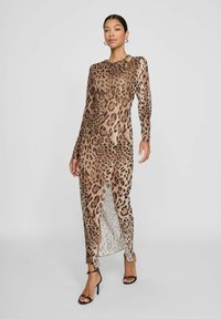 Kvinnlig modell klädd i en lång klänning med leopardmönster och långa ärmar, med en slits fram, tillsammans med svarta högklackade sandaler, mot en enfärgad bakgrund.