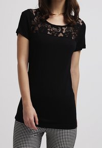 Kvinna iklädd en kortärmad svart topp med spetsdetaljer över bröstet och svart-vita hounds-tooth-byxor.