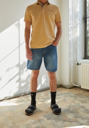 Beige lyhythihainen polo-paita, farkkushortsit, mustat sukat ja mustat sandalit remmeillä, seisoo tekstuuripinnalla luonnonvalossa.