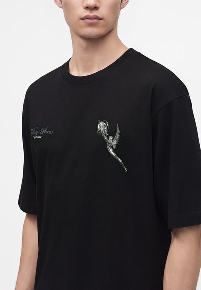 Άνδρας που φοράει μαύρο T-shirt με ασημένια φιγούρα με φτερά που κρατά μια σφαίρα στο αριστερό στήθος και μικρό κείμενο στο δεξί στήθος.