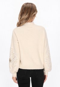 Jasnobeżowy sweter z wysokim dekoltem i dekoracyjnym haftem kwiatowym na rękawach, z koralikami i miękką, teksturowaną powierzchnią. Czarne dżinsy niewidoczne.