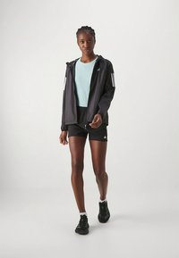 Giacca atletica nera con strisce bianche, top azzurro, pantaloncini e scarpe da ginnastica nere, con chiusura a zip e vestibilità sportiva.