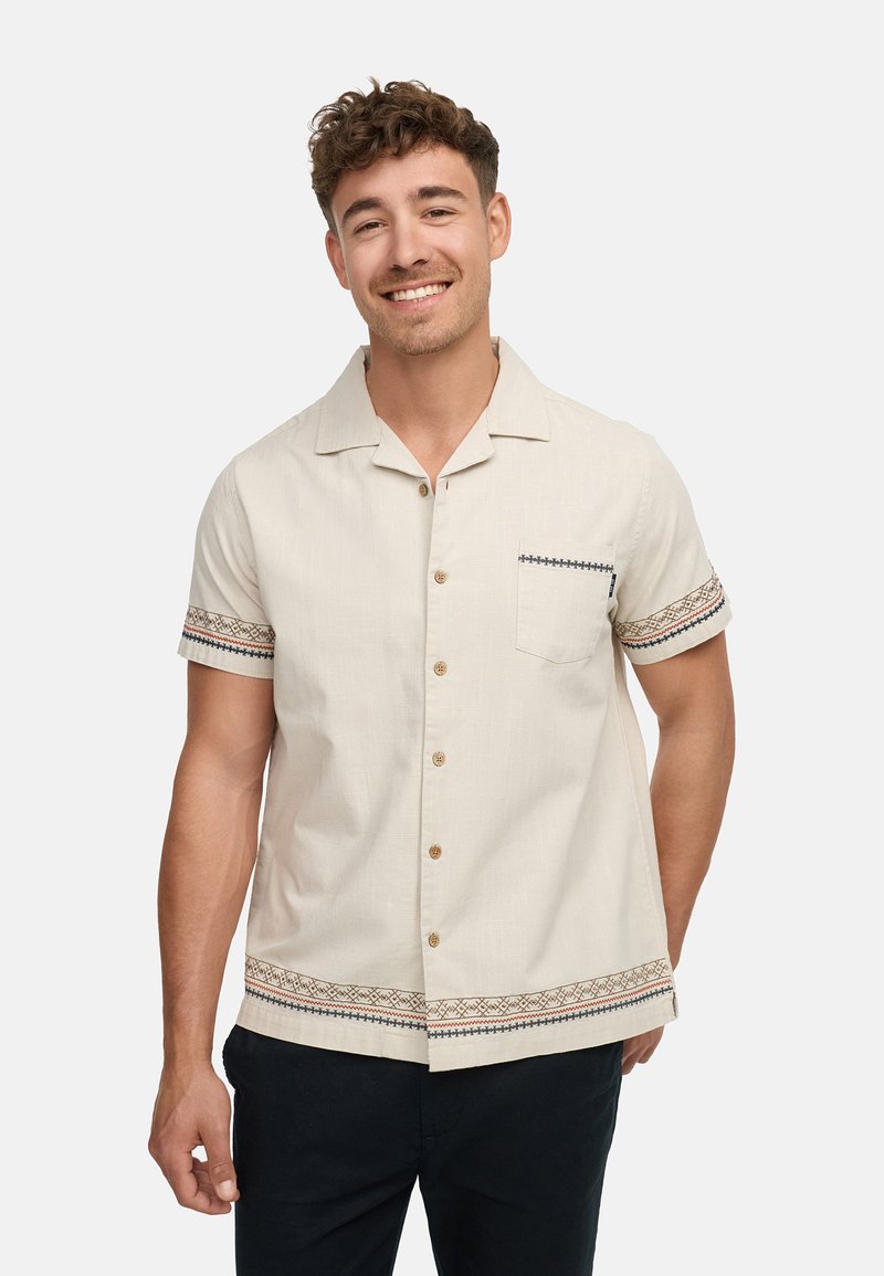 Korte mouwen beige shirt gemaakt van lichte stof, met decoratieve geweven accenten op de manchetten en zoom, een borstzak en knoopsluiting.