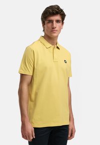 Gul piké med korta ärmar, knappad krage och en liten logotyp på vänster bröst. Tillverkad av mjukt tyg med en slät textur.