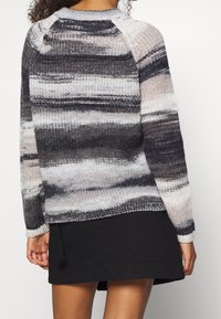 Frau trägt einen schwarzen Rock und einen langärmligen, gestrickten Pullover mit grauen, weißen und schwarzen horizontalen Streifen, von hinten gezeigt.