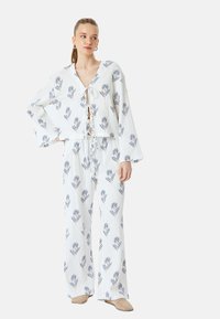 Weißes Stoffoutfit mit blauen Blumenmuster, das aus einem gekürzten Oberteil mit V-Ausschnitt und Glockenärmeln besteht, kombiniert mit weiten Hosen.
