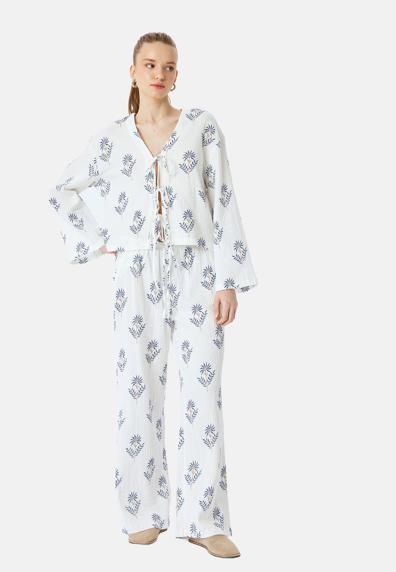 Weißes Stoffoutfit mit blauen Blumenmuster, das aus einem gekürzten Oberteil mit V-Ausschnitt und Glockenärmeln besteht, kombiniert mit weiten Hosen.