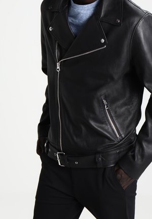 Personne portant une veste de motard en cuir noir avec des poches zippées et une ceinture, associée à une chemise grise et un pantalon noir, les mains dans les poches.