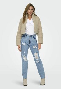 Giacca in pile beige cropped con colletto alto, abbinata a jeans azzurri chiari strappati con diverse lacerazioni. Stivali beige alla caviglia completano l'outfit.