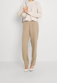 Světle béžový svetr s pleteným vzorem a kulatým výstřihem, v kombinaci s volnými béžovými kalhotami a bílými botami s špičatou špičkou, drží malou kabelku.