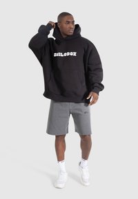 Black hooded sweatshirt with white logo, front pocket, and long sleeves; paired with gray shorts and white sneakers. Neutral background.