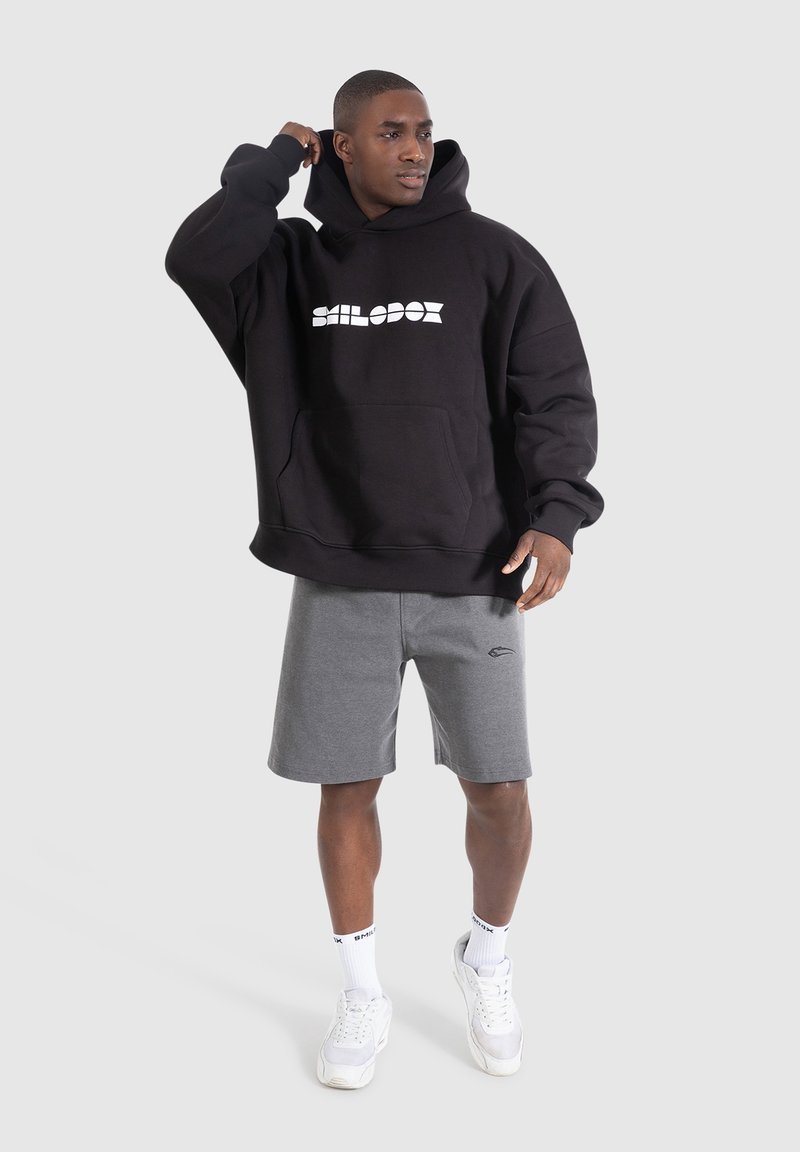 Black hooded sweatshirt with white logo, front pocket, and long sleeves; paired with gray shorts and white sneakers. Neutral background.