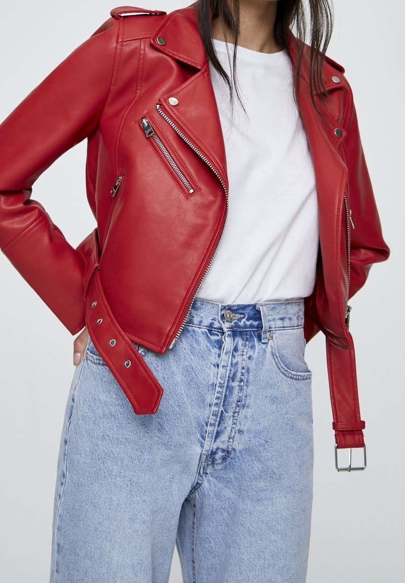 Femme portant une veste de motard en cuir rouge vif par-dessus un T-shirt blanc, associée à un jean denim bleu clair taille haute sur un fond uni.
