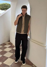 Młody mężczyzna ubrany w beżową koszulkę polo, oliwkowo zieloną pikowaną kamizelkę, czarne spodnie i czarne trampki, stojący wewnątrz przy białej, łukowatej ścianie.