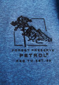 Il tessuto di un blu melangiato presenta un motivo ricamato in nero raffigurante un albero e montagne, con la scritta "FOREST PRESERVE" e "PETROL" in grassetto sottostante.