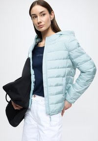 Ανοιχτό μπλε μπουφάν τύπου puffer με φερμουάρ μπροστά και κουκούλα. Διαθέτει κοντινές ραφές και απαλό υλικό. Φορεμένο πάνω από ένα σκούρο μπλε πουκάμισο με λευκό παντελόνι.