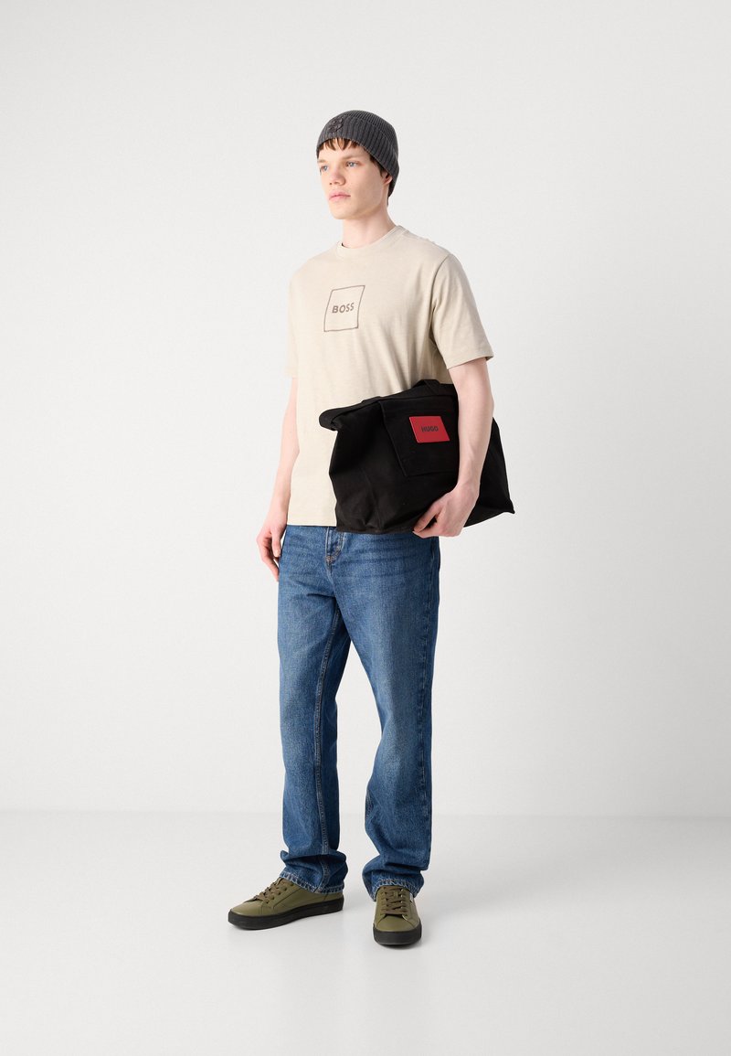Beżowy t-shirt z kwadratowym logo, niebieskie dżinsy, zielono-czarne trampki, dzianinowa szara czapka i czarna torba z czerwonym logo.